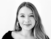 Schwarz-weiße Porträtaufnahme einer lächelnden Person mit langen Haaren und Ohrringen, vor einem weißen Hintergrund.
