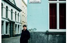 Download Foto: Ein Mann in dunkler Kleidung steht auf einer gepflasterten Straße vor einem hellblauen Gebäude mit roten Vorhängen.