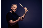 Download Foto: Ein lächelnder Mann in einem schwarzen T-Shirt hält zwei Drumsticks vor einem dunklen Hintergrund hoch.