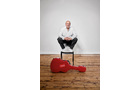 Download Foto: Ein Mann sitzt auf einem Stuhl vor einer weißen Wand und einem Holzboden. Neben ihm liegt ein rotes Gitarrenkoffer.