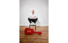 Download Foto: Eine Person sitzt auf einem Stuhl in einem weißen Raum. Vor ihr liegt ein rotes Gitarrencase auf Holzboden.