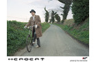 Download Foto: Eine Person im Trenchcoat schiebt ein Fahrrad eine ländliche Straße entlang, umgeben von grünen Hecken und Bäumen.