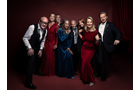 Download Foto: Eine Gruppe von acht elegant gekleideten Personen posiert lächelnd vor einem dunkelroten Hintergrund.