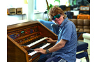 Download Foto: Eine Person in Sonnenbrille spielt eine elektronische Orgel und lächelt. Im Hintergrund sind Möbel und Dekor zu sehen.