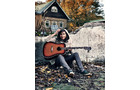 Download Foto: Frau sitzt auf Steinen, hält eine akustische Gitarre. Im Hintergrund ein Holzhaus und herbstliche Bäume.