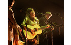 Download Foto: Drei Personen auf einer Bühne: Eine Frau spielt Gitarre und singt, während zwei Männer Instrumente spielen. Bühne in warmem Licht.