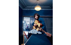Download Foto: Eine Person sitzt in einem blauen Raum auf einem Tisch und spielt Gitarre. Eine Lampe hängt über ihr.