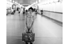 Download Foto: Eine Frau steht mit einem alten Koffer in einer langen, leeren U-Bahn-Passage. Sie trägt ein gemustertes Kleid.