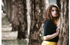 Download Foto: Eine Frau mit langen Haaren lehnt an einem Baumstamm. Sie trägt ein schwarzes Oberteil und einen gelben Rock. Im Hintergrund stehen weitere Bäume.