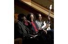 Download Foto: Drei Personen sitzen auf einer Couch. Eine Person hält eine weiße E-Gitarre. Alle lächeln. Hintergrund ist ein gedämpft beleuchteter Raum.