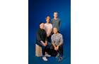 Download Foto: Vier Personen posieren vor einem blauen Hintergrund. Zwei sitzen, zwei stehen dahinter. Alle lächeln entspannt in die Kamera.