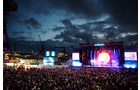 Download Foto: Großes Open-Air-Konzert bei Dämmerung. Menschenmenge vor Bühne mit leuchtenden Bildschirmen und Lichtern, dunkler bewölkter Himmel.
