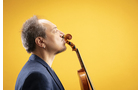 Download Foto: Ein Mann im Profil hält eine Violine, deren Korpus seine Nase berührt, vor einem gelben Hintergrund. Er trägt ein dunkles Sakko.