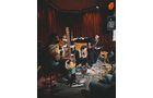 Download Foto: Eine Gruppe von Musikern spielt in einem Raum mit roten Vorhängen, umgeben von Instrumenten wie Gitarren und Trommeln.