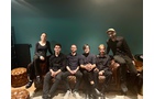 Download Foto: Sechs Personen sitzen nebeneinander auf einem Sofa vor einer grünen Wand, alle in dunkler Kleidung.