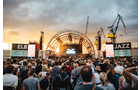 Download Foto: Menschenmenge vor einer beleuchteten Open-Air-Bühne bei Sonnenuntergang, zwei Werbeschilder an den Seiten, Kran im Hintergrund.