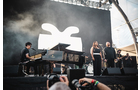 Download Foto: Eine Band tritt auf einer Bühne auf, mit einem großen, abstrakten Logo im Hintergrund. Ein Pianist und mehrere Sänger sind sichtbar.