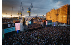 Download Foto: Menschenmenge vor einer großen Open-Air-Bühne mit Kränen im Hintergrund, bei Sonnenuntergang. Gebäude rechts im Bild.