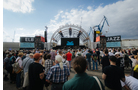 Download Foto: Menschenmenge vor einer Konzertbühne im Freien, mit Jazz-Bannern und Kränen im Hintergrund unter bewölktem Himmel.