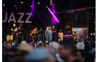 Download Foto: Eine Band performt auf einer Bühne bei einem Jazzfestival, umgeben von bunten Lichtern und einem Publikum im Vordergrund.