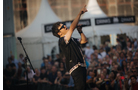 Download Foto: Ein Sänger auf einer Bühne, trägt Sonnenbrille und singt ins Mikrofon, während er nach oben zeigt. Eine große Menschenmenge im Hintergrund.