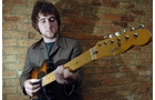 Download Foto: Ein Mann in einer braunen Jacke spielt eine E-Gitarre vor einer Backsteinmauer.