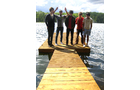 Download Foto: Fünf Personen stehen auf einem Holzsteg am Wasser, eine Person winkt. Im Hintergrund ist ein bewaldetes Ufer zu sehen.