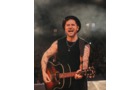 Download Foto: Ein lächelnder Musiker spielt Gitarre auf der Bühne. Er trägt ein schwarzes Oberteil und einen Hut, Tattoos auf den Armen.