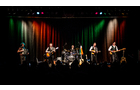 Download Foto: Eine Band spielt auf einer Bühne mit bunten Lichtern im Hintergrund. Fünf Musiker stehen in einer Reihe, Instrumente in der Hand.
