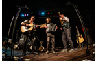 Download Foto: Drei Musiker spielen auf einer Bühne: einer mit Gitarre, einer mit Akkordeon und einer mit Bass. Bunte Lichter im Hintergrund.