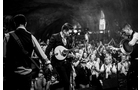 Download Foto: Eine Band spielt in einem voll besetzten, stimmungsvollen Saal. Die Mitglieder tragen Anzüge, und das Publikum klatscht begeistert.