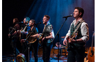 Download Foto: Eine Band mit fünf Musikern spielt auf einer Bühne, jeder mit einem Instrument. Blauer Bühnenhintergrund und Mikrofone sichtbar.