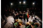 Download Foto: Eine Band steht auf einer Bühne und verbeugt sich vor dem Publikum, im Vordergrund sind Musikinstrumente zu sehen.