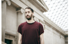 Download Foto: Ein Mann mit Bart steht in einem Gebäude mit heller Steinarchitektur und gläsernem Dach, trägt ein weinrotes T-Shirt.