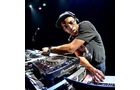Download Foto: Ein DJ mit Kopfhörern steht konzentriert an einem DJ-Pult, die Hand auf einem Mischpult, in einer beleuchteten Bühnenumgebung.