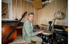 Download Foto: Ein Mann spielt auf einer Trommel in einem Musikraum mit Holzwänden, umgeben von verschiedenen Musikinstrumenten.