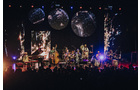 Download Foto: Eine Band spielt auf einer Bühne vor einem Publikum, mit bunten Lichtern und dekorativen Kreisen im Hintergrund.