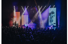 Download Foto: Konzertszene mit bunten Lichtern; eine Band spielt vor einem begeisterten Publikum unter leuchtenden Farbprojektionen.