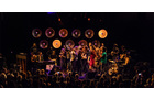 Download Foto: Eine Band und ein Chor performen auf einer Bühne mit buntem Licht und großen, runden Lautsprechern im Hintergrund vor Publikum.