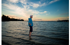 Download Foto: Eine Person steht in flachem Wasser, blickt in die Ferne. Der Himmel ist teils bewölkt bei Sonnenuntergang.