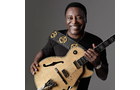 Download Foto: Eine Person lächelt und hält eine E-Gitarre. Sie trägt ein dunkles T-Shirt mit gelbem Muster vor grauem Hintergrund.