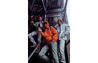 Download Foto: Drei Personen stehen auf einer Metalltreppe. Eine trägt eine orangene Jacke, die anderen weiße Kleidung. Sie schauen zur Kamera.