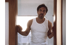 Download Foto: Mann in weißem Tanktop steht in einer Türöffnung, hält die Türrahmen mit beiden Händen fest und schaut nachdenklich.