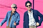 Download Foto: Zwei Personen mit Sonnenbrillen stehen vor einer pinken Ziegelwand. Eine Person trägt eine Jeansjacke, die andere einen dunkelblauen Mantel.