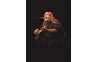 Download Foto: Mann mit langen Haaren spielt Gitarre und singt in ein Mikrofon auf einer dunklen Bühne.