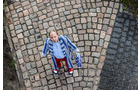 Download Foto: Ein Mensch im blauen gestreiften Mantel steht auf einem Kopfsteinpflaster und blickt nach oben, umgeben von grauen Steinen.