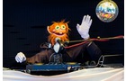Download Foto: Ein orangefarbener Puppen-Charakter mit Handschuhen steht an einem DJ-Pult und winkt. Im Hintergrund hängt eine Discokugel.