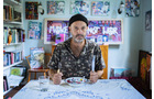 Download Foto: Ein Mann sitzt an einem Tisch mit Besteck, umgeben von bunter Kunst und Büchern in einem kreativen Raum.