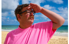 Download Foto: Eine Person steht am Strand und trägt ein pinkes T-Shirt. Sie blickt in die Ferne und hält die Hand über die Augen. Im Hintergrund Meer und Himmel.
