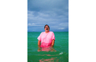 Download Foto: Person in pinkem T-Shirt steht lachend im Meer, der Himmel ist bewölkt.
